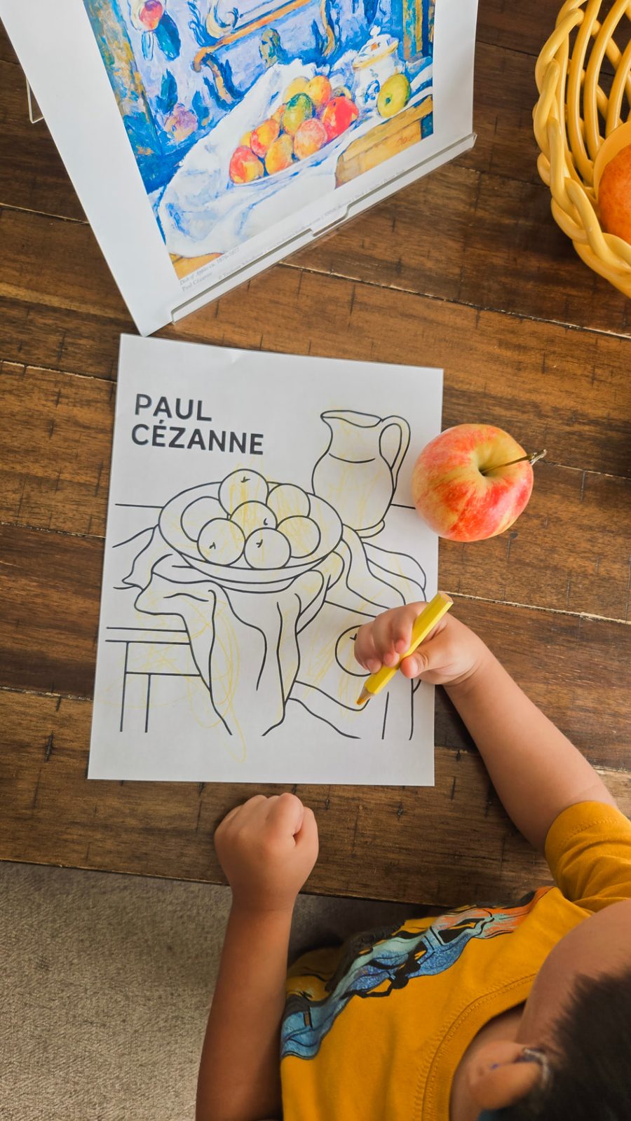A child coloring a Paul Cézanne still-life page at a wooden table, with the art print and a real apple beside the paper